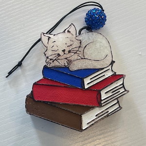 May include: A white cat figurine sleeping on a stack of three books. The books are brown, red, and blue. A blue sparkly ornament hangs from the top of the figurine.