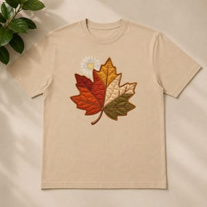 May include: A beige short-sleeved t-shirt featuring a large, quilted-style maple leaf graphic on the chest. The leaf is divided into sections of red, orange, yellow, cream, and olive green, with a small white daisy flower positioned near the top.