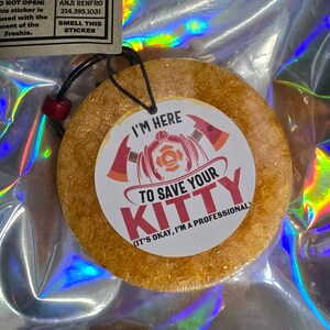 May include: A round, gold-colored car air freshener with a white sticker that says "I'm here to save your kitty (It's okay, I'm a professional)". The sticker features a cartoon firefighter with a fire hose and an axe.