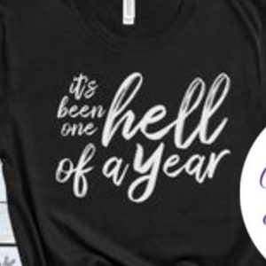 May include: Black t-shirt with white text that reads "It's been one hell of a year". The shirt is knotted at the bottom. The image also includes a watermark that reads "Cool Tees & Freshies".