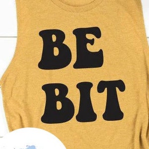 May include: A mustard yellow sleeveless tank top with the text "BE BIT" in black retro-style lettering.
