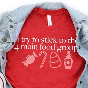 May include: Red t-shirt with the text "I try to stick to the 4 main food groups" and a graphic of four candy icons. The shirt is folded and laid on a white surface with a denim jacket and white sneakers.