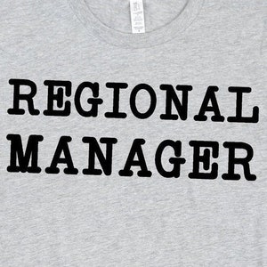 May include: A gray t-shirt with the text "REGIONAL MANAGER" printed in black. The shirt is displayed on a white background with a pair of black sneakers and blue jeans.