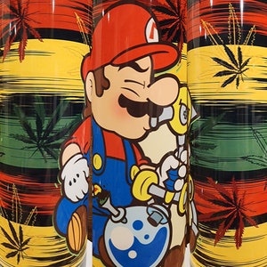 May include: A stainless steel tumbler with a colorful design featuring a cartoon character holding a bong. The design includes a red, yellow, green, and black color scheme with a marijuana leaf pattern.