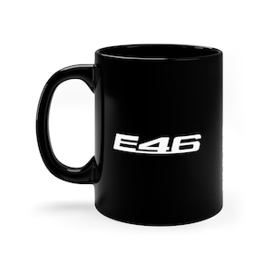 May include: A black ceramic mug with a white "E46" logo. The mug has a curved handle and a cylindrical shape. The logo is centered on the mug's surface.