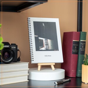 Może przedstawiać: Biały notes w spirali z czarno-białym zdjęciem budynku na okładce. Notes stoi na małym drewnianym sztalugach i znajduje się na okrągłym białym podkładzie. Na okładce widnieje napis "Daily Notes".