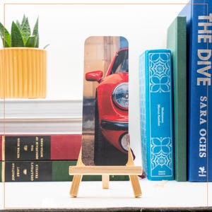 Puede incluir: Un marcapáginas con un coche rojo vintage se muestra en un pequeño caballete de madera. El marcapáginas se coloca frente a una pila de libros y una planta suculenta en maceta. El faro y el espejo lateral del coche son visibles.