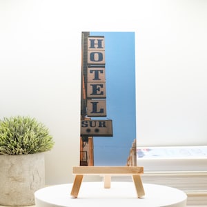 May include: A photograph of a vintage hotel sign with the word "HOTEL" stacked vertically and "SUR" below. The sign is set against a blue sky, mounted on a wooden easel.