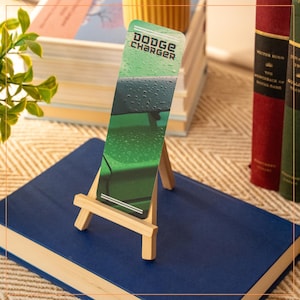 Puede incluir: Un marcapáginas con el texto "Dodge Charger" sobre un caballete de madera, apoyado en un libro azul. El marcapáginas presenta una imagen de coche verde con gotas de agua. Libros en el fondo.