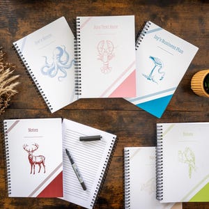 May include: A collection of spiral-bound notebooks with white covers, featuring illustrations of an octopus, lobster, bird, deer, and parrot. Each notebook has a coloured diagonal stripe and text like "Joe's Notes". A pen lies on an open notebook.