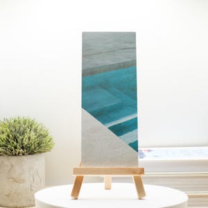 May include: A rectangular art print featuring an abstract design of a pool with turquoise water and gray stone. The print is displayed on a small wooden easel, set against a white background. A small potted plant and books are also visible.