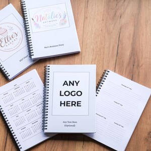 May include: Four spiral-bound notebooks with white covers. One features a colourful Natalie's Artwork logo, another shows an Ellie's Bakery logo, and a third displays the black text ANY LOGO HERE with space for optional text below. One notebook is open.