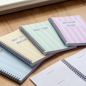 May include: Four spiral-bound notebooks with striped covers in light blue, yellow, green, and pink. The covers feature white rectangular labels with black text reading Emily's 2021 Work Diary, Rachel's Diary, and Any Custom Text Here.