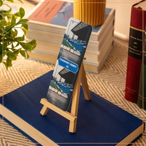 Puede incluir: Un marcapáginas azul y blanco con una foto de un estadio y el texto "Stockport County FC". El marcapáginas está sobre un caballete de madera.