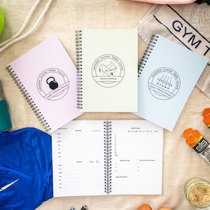 Könnte beinhalten: Eine Sammlung von Fitness-Journals in verschiedenen Farben, darunter Rosa, Hellgrün und Blau, mit Spiralbindung. Jedes Journal hat ein rundes Logo mit Text zu Ernährung, Bewegung, Gewohnheiten und Wachstum. Ein Journal ist geöffnet und zeigt linierte Seiten.