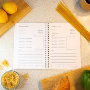 Puede incluir: Un libro de recetas abierto con páginas en blanco para escribir recetas. El libro tiene una encuadernación en espiral y está sobre una superficie blanca con varios ingredientes de cocina alrededor, incluyendo limones, espaguetis, arroz, boniatos y un pimiento verde.