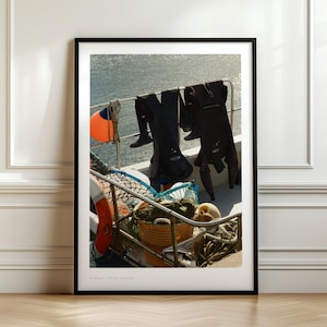 May include: A framed photograph featuring diving gear drying on a boat. Black wetsuits hang from a railing, with an orange buoy and a life preserver visible. A wicker basket and fishing nets are in the foreground, with the ocean in the background.