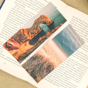 May include: Two rectangular bookmarks with scenic images. One shows a coastal rock formation with an arch, the other a beach with tall grass. The bookmarks are on a book page with text.