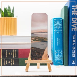 May include: A bookmark featuring a scenic landscape photograph of a small island with a body of water and a house in the distance. The bookmark is displayed on a wooden easel.