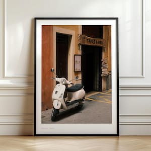 May include: Framed print of a white scooter parked in front of a restaurant entrance. The restaurant has a wooden sign that reads "TAPAS & BBQ". The image has a warm, inviting color palette, with the scooter as the focal point.