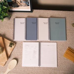May include: Four spiral-bound recipe books in white, blue, and green. The open book shows recipe fields and lined pages. Each cover reads "RECIPE BOOK". A wooden chopping board, spoon, and pasta are also visible.