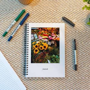 Puede incluir: Un cuaderno blanco de espiral con una foto colorida de un mercado de flores en la portada. La foto muestra una variedad de flores, incluyendo girasoles, rosas y lirios. El texto "Journal" está impreso en negro en la parte inferior de la portada.