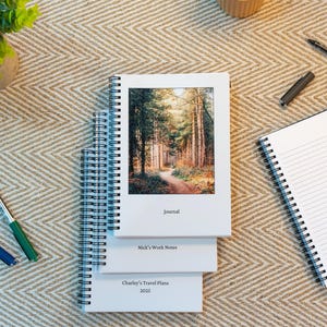 May include: Three white spiral bound notebooks stacked on top of each other. The top notebook has a photo of a forest path on the front cover and the text "Journal". The middle notebook has the text "Nick's Work Notes" and the bottom notebook has the text "Charley's Travel Plans 2025".