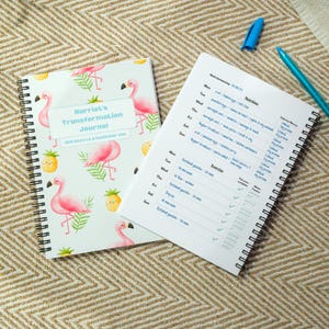 May include: A journal with a flamingo and pineapple design, spiral-bound. The cover says "Harriet's Transformation Journal" and "365 days to a healthier you." An open page shows a nutrition and exercise tracker, with a blue pen and marker.