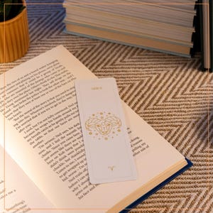May include: A white bookmark featuring a gold outline of an Aries zodiac symbol and stars. The bookmark is labelled "Aries" at the top and has the symbol for Aries at the bottom.