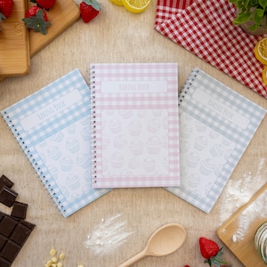 May include: Three spiral-bound "Baking Book" notebooks in pastel shades: blue, pink, and grey. Each notebook has a gingham pattern border and a white label with the title. The centre of each notebook has a cupcake pattern.