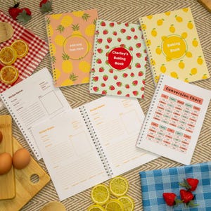 Puede incluir: Una colección de libros de recetas con espiral y cubiertas coloridas con diseños de piñas, fresas y limones. Un libro dice "Charley's Baking Book", y otro dice "Baking Book". También se ven páginas de recetas abiertas y una tabla de conversión.