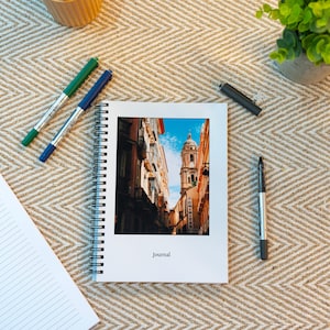 May include: A white spiral bound journal with a photo of a narrow street in a European city. The photo shows a tall building with a clock tower and a blue sky. The journal has the word "Journal" printed on the front cover.
