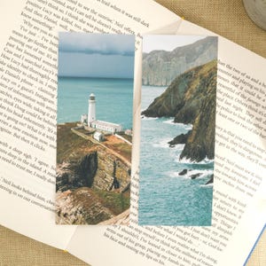 May include: Two rectangular bookmarks featuring coastal landscape photography. One shows a white lighthouse on a cliff overlooking the ocean. The other depicts a rocky coastline with waves crashing against the shore. Both are set against the backdrop of an open book.