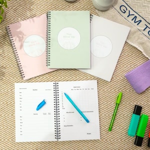 Könnte beinhalten: Spiralgebundene Notizbücher in Pastellfarben, Rosa, Grün und Weiß, mit dem Text "The Glow Up" auf dem Cover. Ein offenes Notizbuch zeigt linierte Seiten mit einem blauen Stift, sowie grüne und türkisfarbene Stifte und Textmarker.