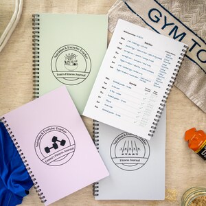 Pode incluir: Uma coleção de diários de fitness com espiral em várias cores, incluindo verde claro, rosa e branco. Cada diário apresenta um logotipo circular com texto como "Nutrition & Exercise Tracker" e "Fitness Journal". Um diário está aberto, mostrando um plano semanal de exercícios e nutrição.