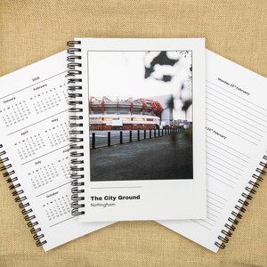 May include: A white spiral-bound notebook featuring a photograph of 'The City Ground Nottingham'. Calendars for 2026 and daily planner pages are visible. The image is set against a neutral background.