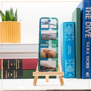 May include: A rectangular bookmark with images of the Scottish Highlands, including a signpost, landscape, and a Highland cow. The text "THE HIGHLANDS" is printed on the bookmark. It is displayed on a small wooden easel, with books and a potted plant in the background.