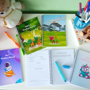 Op de afbeelding: Een verzameling kleurrijke spiraalgebonden leesdagboeken met verschillende omslagontwerpen, waaronder een astronaut, een dinosaurus en een eenhoorn. De dagboeken zijn open en tonen gelinieerde pagina's. Pennen en stiften liggen erbij.
