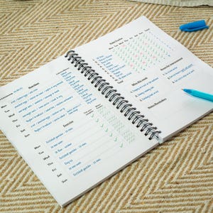 Peut inclure: Un agenda à spirales ouvert, avec des sections pour la nutrition, l'exercice et le suivi des habitudes. Les pages blanches comportent du texte bleu et des cases à cocher. Un stylo bleu est posé sur la page, sur un fond beige.