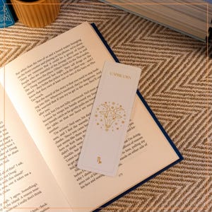 Könnte beinhalten: Ein weißes Lesezeichen mit einer goldenen Illustration eines Steinbock-Sternzeichen. Das Lesezeichen hat den Schriftzug "Steinbock" in goldenen Buchstaben oben. Das Lesezeichen liegt auf einem geöffneten Buch.