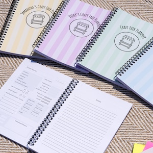 May include: Spiral-bound craft fair planners with pastel striped covers. Each planner features a different color scheme and the text "Craft Fair Planner" with a market stall illustration. One planner is open, revealing lined pages.