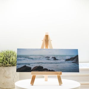 May include: A rectangular print of a seascape featuring crashing waves and dark rocks. The print is displayed on a small wooden easel. The scene is dominated by shades of blue and gray, with a cloudy sky and a hint of green vegetation.