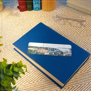 May include: A blue hardback book with a silver bookmark featuring a photo of a stadium. The bookmark is placed on the book, and the book is lying on a woven surface.
