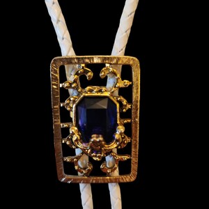 May include: A white bolo tie with a gold-toned pendant featuring a blue gemstone set in a decorative frame.