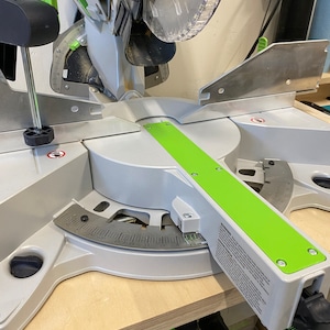 May include: A silver and green miter saw with a clear blade guard. The saw features a green base and a degree scale for precise angle cuts. The saw is designed for woodworking and construction projects.