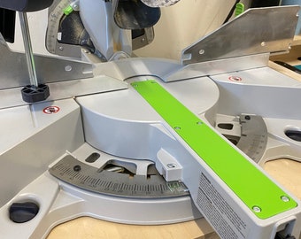 May include: A silver and green mitre saw with a clear blade guard. The saw has a green base and a degree scale for accurate angle cuts. The tool is designed for woodworking and construction projects.