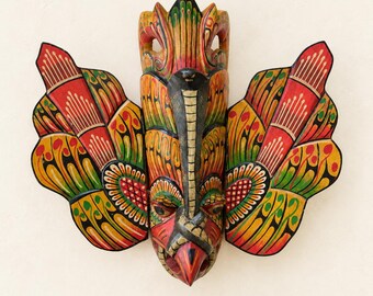 May include: A hand-carved wooden wall mask featuring a central figure with a cobra hood and a bird-like beak. The mask is painted in vibrant red, yellow, green, and black colors with intricate patterns on the two large, wing-like side panels.