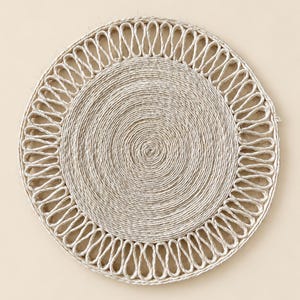 May include: A round, beige placemat crafted from coiled natural fiber rope. The center features a tight, circular spiral pattern, while the outer border consists of a decorative, open-loop design created by looping the same light-colored cord.