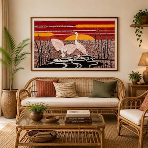 Op de afbeelding: Een rechthoekige wandkunstprint met twee witte kraanvogels die in een donkere vijver met hoog riet staan, tegen een levendige achtergrond van rode en oranje horizontale banden en een gele zon, geplaatst in een eenvoudige houten lijst.