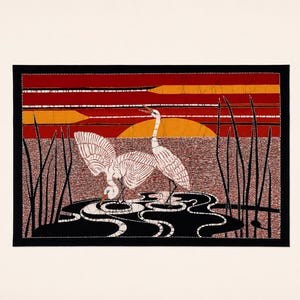 May include: A rectangular textile art piece featuring two white cranes in a dark pond with black reeds. The background displays a sunset with horizontal bands of red and orange. The scene is framed by a solid black border on a cream-colored mat.